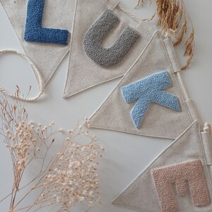 May include: A banner with the name "LUKE" spelled out in colorful yarn letters. The letters are sewn onto triangular fabric flags. The flags are connected by a white string.