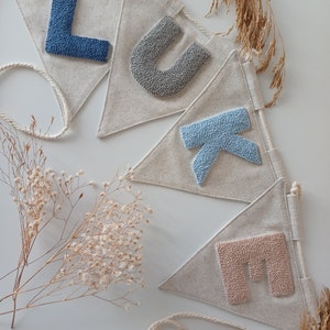 May include: A banner with the name "LUKE" spelled out in felt letters. The letters are in shades of blue and gray. The banner is made of natural linen fabric and is strung with twine.
