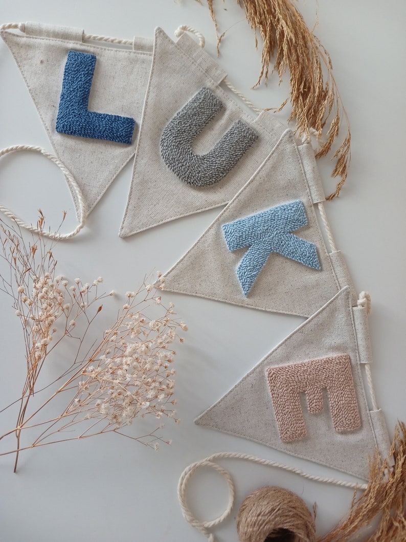 May include: A banner with the name "LUKE" spelled out in felt letters. The letters are in shades of blue and gray. The banner is made of natural linen fabric and is strung with twine.
