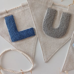 May include: A banner with the letters "LU" in blue and gray yarn, sewn onto a natural linen fabric. The banner is strung with a white rope.