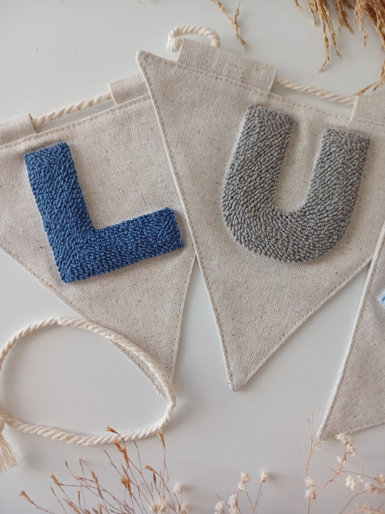 May include: A banner with the letters "LU" in blue and gray yarn, sewn onto a natural linen fabric. The banner is strung with a white rope.