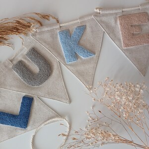 May include: A banner with the name "LUKE" spelled out in felt letters. The letters are in different colors: gray, blue, light blue, and pink. The banner is made of natural linen fabric and is strung together with a white cord.