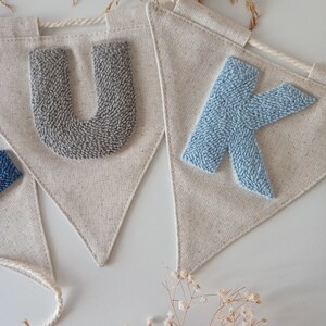May include: A banner with the letters "UK" in gray and blue yarn, sewn onto a natural linen fabric. The banner is strung with white twine.