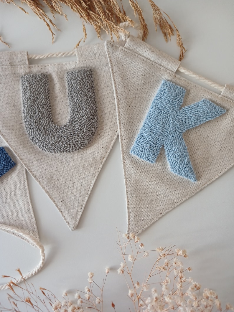 May include: A banner with the letters "UK" in gray and blue yarn, sewn onto a natural linen fabric. The banner is strung with white twine.