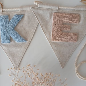May include: A light blue "K" and a light pink "E" are stitched onto a beige fabric banner. The banner is made of a natural linen material and has a frayed edge. The letters are made of yarn and are raised from the fabric.