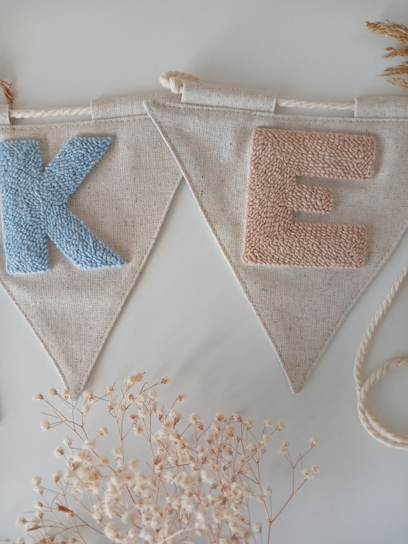 May include: A light blue "K" and a light pink "E" are stitched onto a beige fabric banner. The banner is made of a natural linen material and has a frayed edge. The letters are made of yarn and are raised from the fabric.