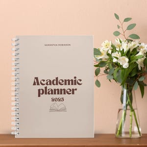 May include: A beige academic planner with a brown title that reads "Academic Planner 2025" and a small illustration of a book. The planner is on a wooden surface with a vase of white flowers in the background.