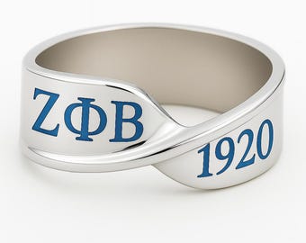 Pode incluir: Um anel prateado com as letras azuis "ZΦB" e o número "1920". O anel tem um design curvo e polido. O interior do anel é de uma cor prateada mais clara. É provavelmente uma joia.