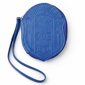 May include: Blue round coin purse with a shield design. The purse has a wrist strap and a zipper closure. The shield design includes the letters "ZOB" and the year "1920". The purse is made of a textured material.