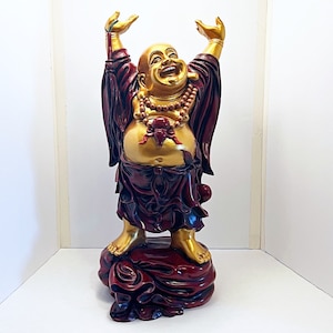 May include: A golden laughing Buddha statue with raised arms, wearing a red robe with gold accents. The statue is standing on a red base with a flowing fabric design.