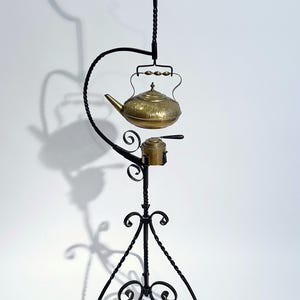 May include: A brass teapot with an ornate design hangs from a black wrought iron stand with a twisted base. The stand has a decorative scrollwork design.