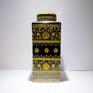 Op de afbeelding: Een zwarte en gouden keramische pot met een vierkant lichaam en een deksel. De pot is versierd met ingewikkelde bloemmotieven en geometrische ontwerpen.