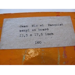 Może przedstawiać: Etykieta z napisem "Jean Michel Basquiat acryl on board 34,3 x 44,5 cm 160" jest przymocowana do pomarańczowej powierzchni. Etykieta jest prostokątna i wykonana z jasnego, teksturowanego papieru.