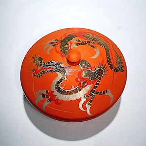 Puede incluir: Contenedor redondo naranja con tapa decorada con un diseño de dragón en negro, plateado y rojo. La tapa tiene un pequeño pomo redondo. El contenedor es probablemente de cerámica o madera lacada.