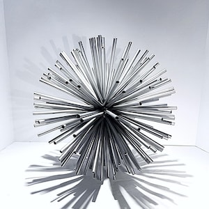 May include: A contemporary, spherical sculpture composed of many polished, silver metal tubes extending outwards. The sculpture casts a strong shadow on the white surface. This decorative piece is ideal for modern home decor. The sculpture's diameter is approximately 25 cm.
