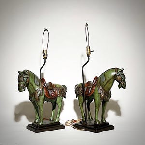 May include: Two green horse-shaped table lamps with brown saddles and decorative accents. The lamps have black stems and gold-colored bulb sockets. The lamps are on dark brown rectangular bases.
