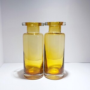 Op de afbeelding: Twee amberkleurige glazen flessen met een heldere glazen bovenkant en onderkant. De flessen zijn cilindrisch en hebben een kleurverloop van licht naar donker amber.