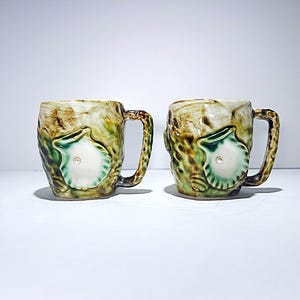 Puede incluir: Dos tazas de cerámica con un diseño texturizado y terroso. Cada taza presenta un motivo de concha marina en relieve en blanco y verde, con un pequeño detalle en forma de perla. Las asas son de un marrón más oscuro, complementando el esquema de color general.