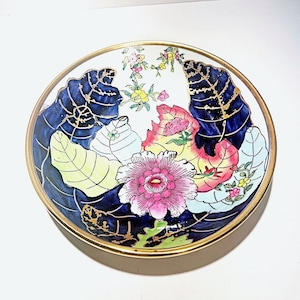 May include: A decorative ceramic plate with a gold rim. The plate features a floral design with pink, yellow, and blue leaves. The center of the plate has a large pink flower.