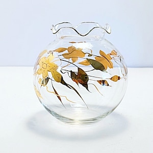 May include: A clear glass vase with a round body and a ruffled rim. The vase is decorated with gold-coloured floral and leaf designs. The flowers are yellow, and the leaves are a mix of gold and green. The vase is a decorative item.