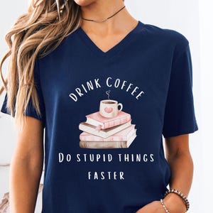 May include: Navy blue V-neck t-shirt with the text "DRINK COFFEE DO STUPID THINGS FASTER". The graphic features a coffee cup on a stack of books. The shirt is a casual wear item.