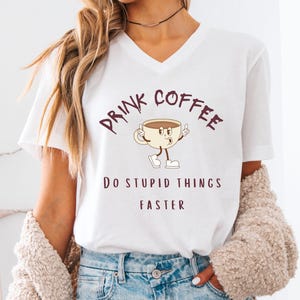 May include: White V-neck t-shirt with the text "DRINK COFFEE DO STUPID THINGS FASTER" in brown lettering. The shirt features a cartoon coffee cup character. The model is wearing light blue denim shorts and a beige cardigan.
