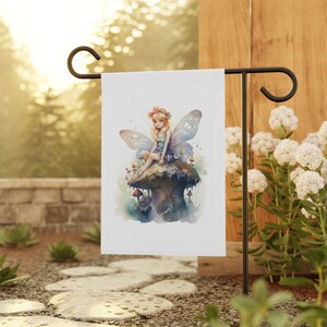 May include: A white garden flag with a watercolor illustration of a fairy sitting on a mushroom. The fairy has blonde hair, blue wings, and is wearing a blue dress. The mushroom is brown and has white spots.