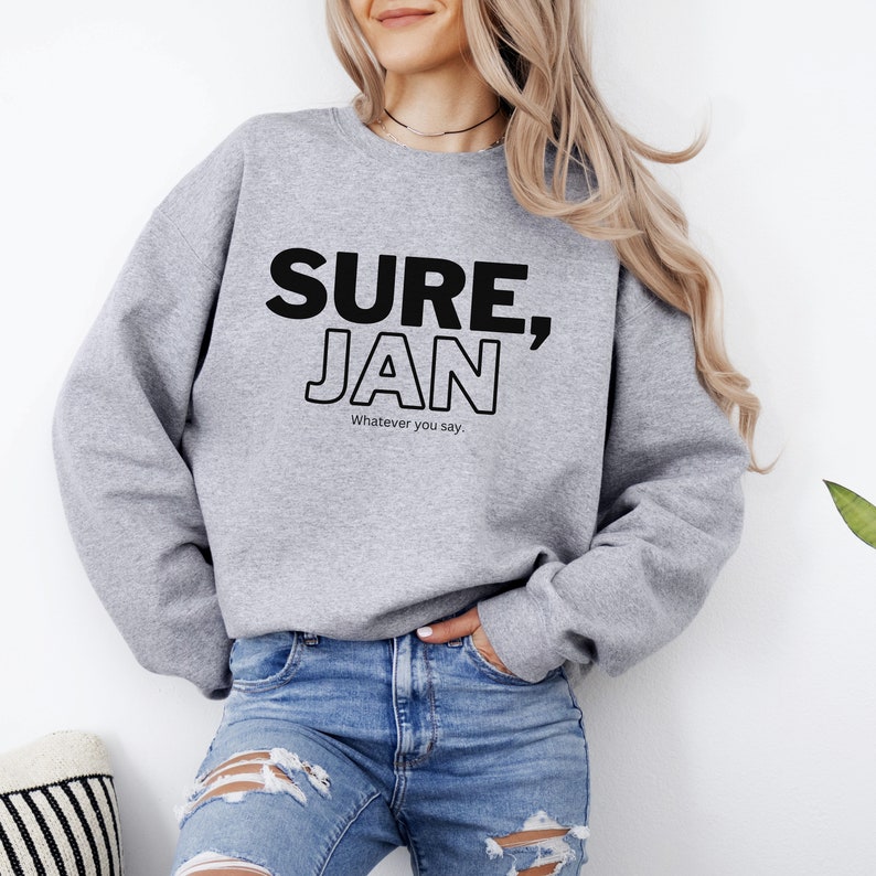 May include: Gray sweatshirt with the text "SURE, JAN" in black outlined letters. The text "Whatever you say." is printed below in smaller black letters.