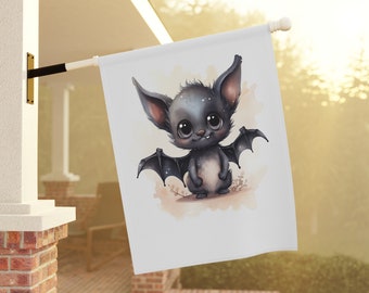May include: A white garden flag features a cute cartoon bat with large eyes and wings. The bat is primarily dark gray with lighter gray accents and is set against a soft, watercolor-style background. The flag is displayed outdoors.
