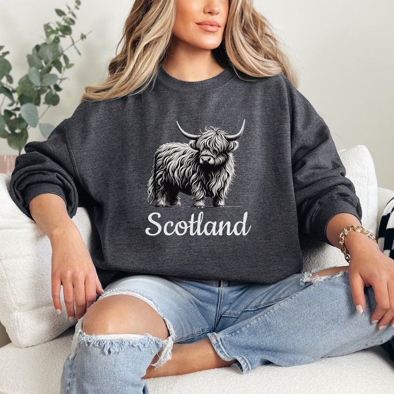 Puede incluir: Una sudadera gris oscuro con un gr&aacute;fico blanco de una vaca de las Highlands y la palabra "Escocia" impresa debajo.