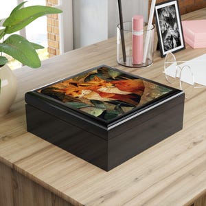 May include: A black wooden box with a hinged lid. The top of the box features a detailed illustration of two foxes nestled among leaves in shades of orange, brown, and green. The box is sitting on a wooden surface.