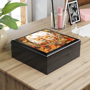 May include: A black wooden box with a hinged lid. The lid features a watercolor painting of two foxes nestled among autumn leaves. The box is rectangular and sits on a wooden surface, suggesting a decorative or storage item.