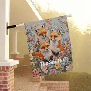 May include: A garden flag with a blue background featuring three orange foxes surrounded by colorful flowers. The text "The Smiths" is printed in white at the bottom of the flag.