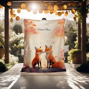 May include: A soft, cozy blanket featuring a watercolor illustration of two foxes in a forest setting. The foxes are facing each other and appear to be kissing. The background is a soft pink and orange with falling hearts. The text "Custom Name" is printed in black at the top of the blanket.