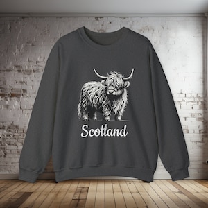 Puede incluir: Una sudadera gris oscuro con un gr&aacute;fico blanco de una vaca de las tierras altas y la palabra "Escocia" impresa debajo.