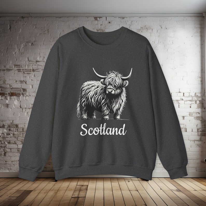 Puede incluir: Una sudadera gris oscuro con un gr&aacute;fico blanco de una vaca de las tierras altas y la palabra "Escocia" impresa debajo.