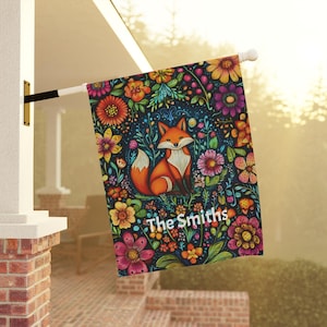 May include: A colorful garden flag with a fox and flowers. The flag has a dark blue background with a variety of flowers in shades of red, orange, yellow, pink, and purple. The fox is orange and white with a bushy tail. The text "The Smiths" is printed in white at the bottom of the flag.