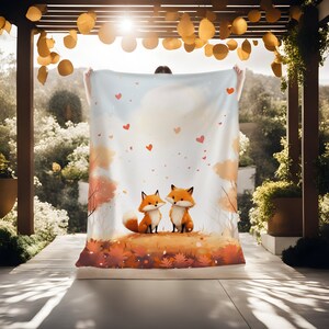 May include: A white throw blanket with a whimsical illustration of two orange foxes with red hearts floating around them. The foxes are standing in a field of autumn leaves.