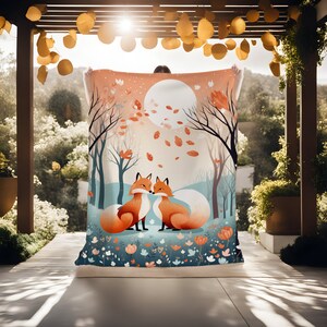 May include: A throw blanket with a whimsical illustration of two foxes in a forest setting. The foxes are orange and white, and they are surrounded by trees, leaves, and flowers. The background is a soft blue and orange.