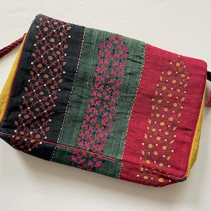 Peut inclure: Une pochette brodée colorée avec une bordure jaune. Le sac est fait d'un tissu foncé avec un motif rayé vert, rouge et noir. Les rayures sont décorées de broderies complexes en rose, jaune et blanc.