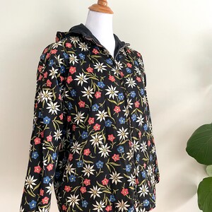 May include: A black hooded jacket with a floral print featuring white, red, and blue flowers. The jacket has a zip closure and a drawstring hood.