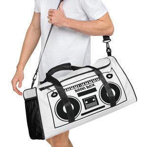 Pode incluir: Bolsa de viagem branca com um design de toca-fitas preto e branco. A bolsa tem um zíper preto e uma alça de ombro preta ajustável.