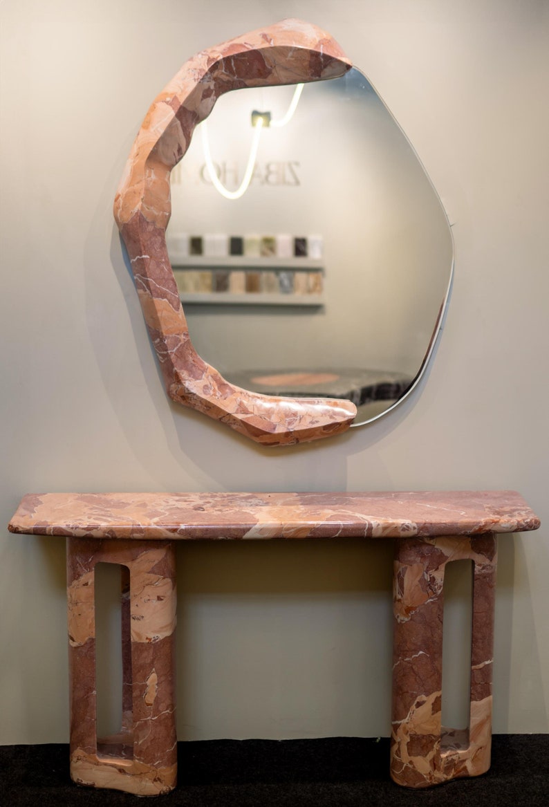May include: A pink marble console table with a unique, organic design. The table has a rectangular top and two legs with openwork details. A large, irregularly shaped mirror with a pink marble frame hangs above the table.