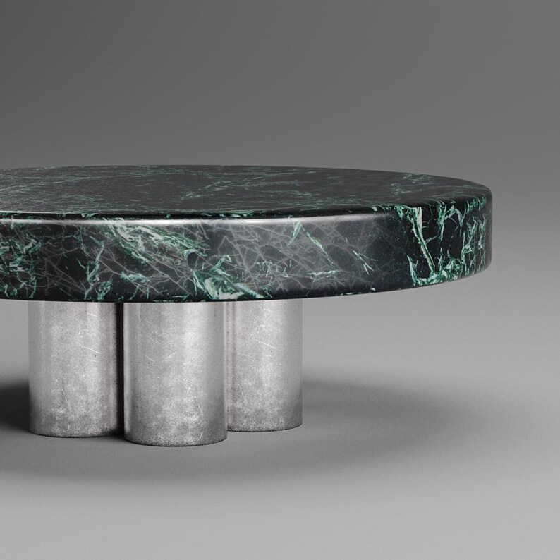 May include: A round coffee table with a dark green marble top and three silver cylindrical legs. The marble has a natural veining pattern in shades of green and white. The table is a modern design.