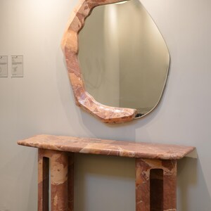 May include: A pink marble console table with a unique, organic design. The table has a rectangular top and two legs with openwork details. A large, irregularly shaped mirror with a pink marble frame hangs above the table.