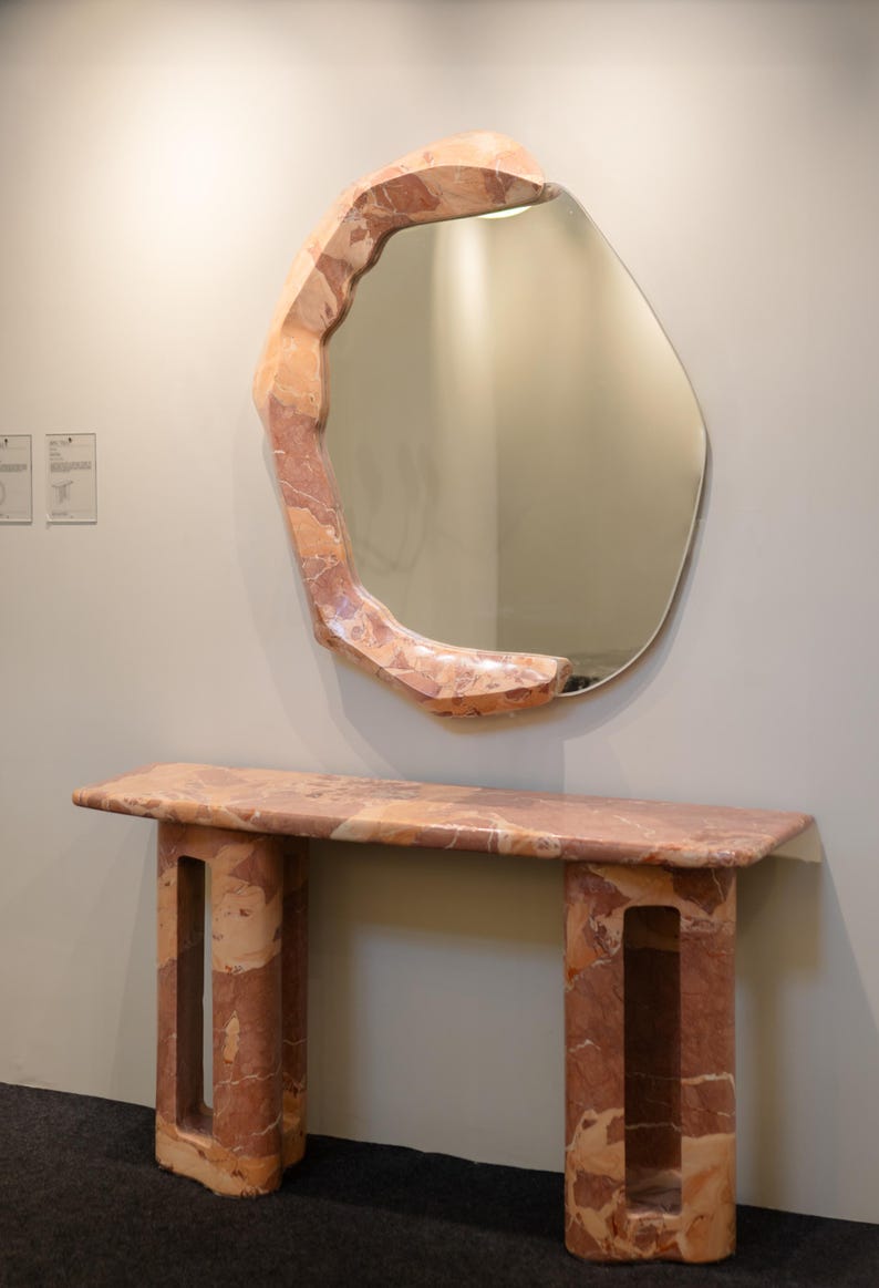 May include: A pink marble console table with a unique, organic design. The table has a rectangular top and two legs with openwork details. A large, irregularly shaped mirror with a pink marble frame hangs above the table.