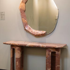 May include: A pink and brown marble console table with a unique, organic shape. The table has two legs with open spaces and a rectangular top. A large, irregularly shaped mirror with a pink and brown marble frame hangs above the table.
