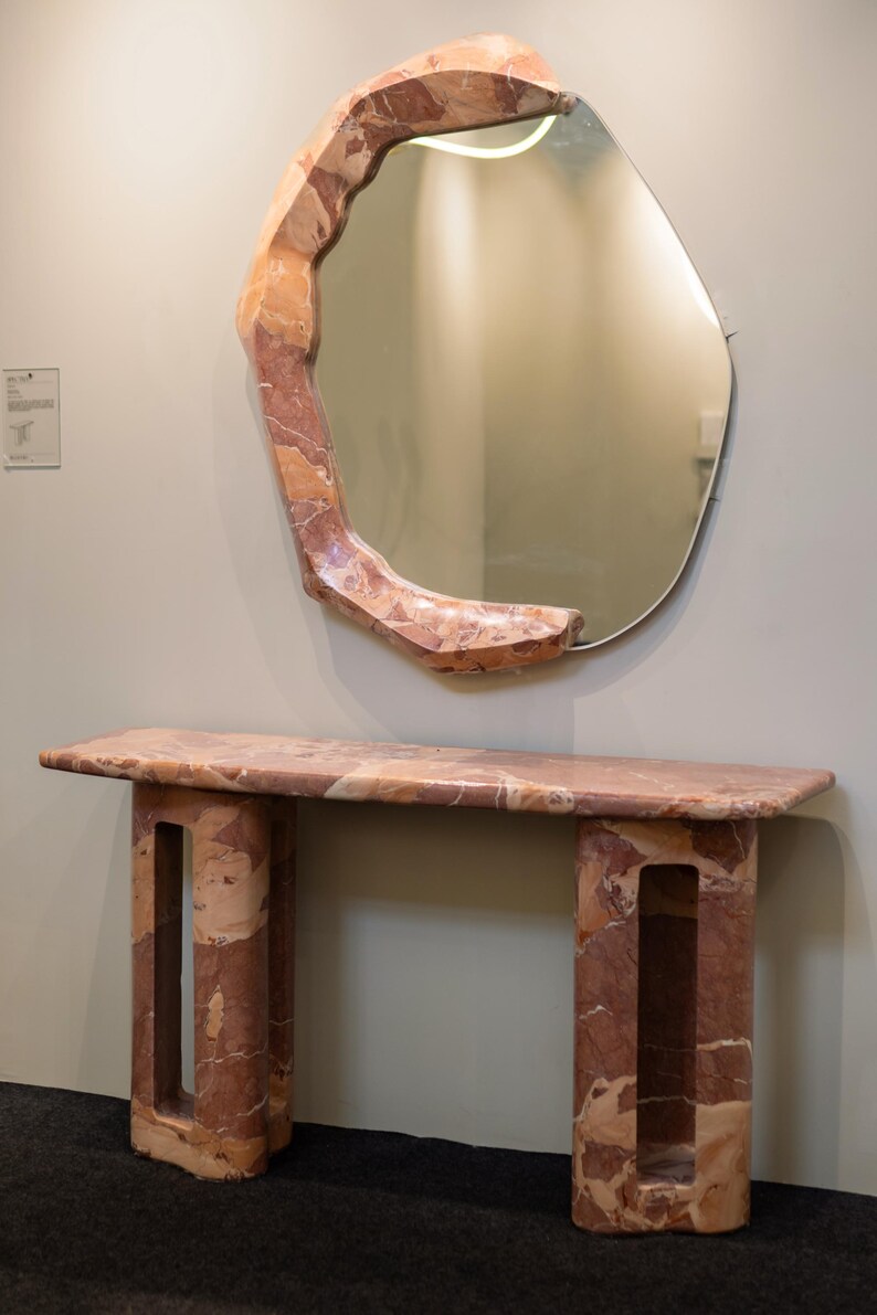 May include: A pink and brown marble console table with a unique, organic shape. The table has two legs with open spaces and a rectangular top. A large, irregularly shaped mirror with a pink and brown marble frame hangs above the table.