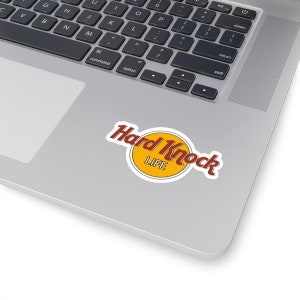 Könnte beinhalten: Ein weißer Aufkleber mit einem roten und gelben Logo, auf dem "Hard Knock Life" steht.