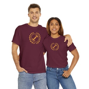 Könnte beinhalten: Zwei Personen tragen weinrote T-Shirts mit einem goldenen Aufdruck eines Buchstabens "I" in einem Kranz. Der Aufdruck hat einen abgenutzten Look.